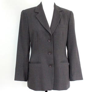 May include: A charcoal gray blazer featuring a notched lapel, long sleeves, and a three-button front closure. The garment includes two horizontal zippered pockets at the waist and a single welt pocket on the upper left chest area.