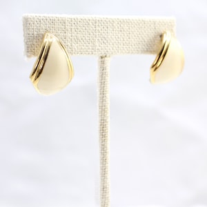 May include: A pair of cream-coloured, triangular earrings with a gold-tone trim. The earrings are displayed on a white background, showcasing their geometric shape and elegant design. The earrings are a classic accessory.