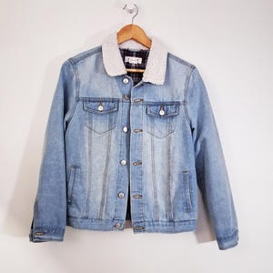 May include: Light blue denim jacket with a cream-colored, fuzzy collar. The jacket has silver buttons, two chest pockets, and two side pockets. The interior lining is a plaid pattern. The jacket is hanging on a wooden hanger.