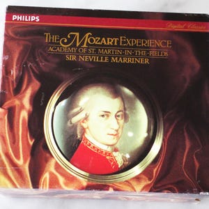 May include: A CD box set titled "The Mozart Experience" by Philips. The box features a portrait of Mozart in a gold-colored frame. The text on the box includes "Academy of St. Martin-in-the-Fields" and "Sir Neville Marriner". The background is a dark, textured pattern.