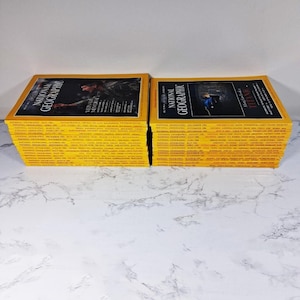 May include: Two stacks of vintage National Geographic magazines. The covers are yellow with black text and images. One stack features a cover with a man, and the other features a cover with a person in a dark room. The magazines are stacked on a white surface.