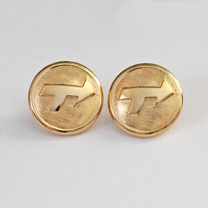 May include: A pair of round gold-tone metal buttons featuring a textured background and a polished, raised arrow symbol pointing toward the right. The circular buttons have a smooth, raised outer rim and a flat, reflective surface finish.