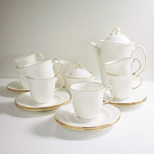May include: A white porcelain tea set with gold edging. The set includes a teapot, sugar bowl, saucers, and teacups. The teacups are stacked, and the set is arranged on a white surface. The gold edging adds a touch of elegance to the tea set.