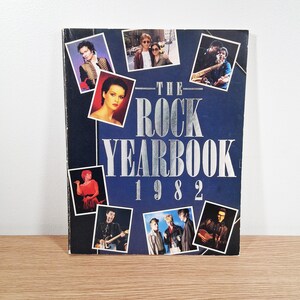 May include: A vintage "The Rock Yearbook 1982" book with a navy blue cover. The cover features photos of musicians and celebrities, with the title in silver lettering. The book is on a wooden surface.