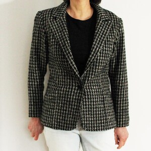 May include: A black and white houndstooth blazer with a notched lapel and a single-button closure. The blazer has a classic pattern and a tailored fit. The person is wearing a black shirt and light blue jeans.