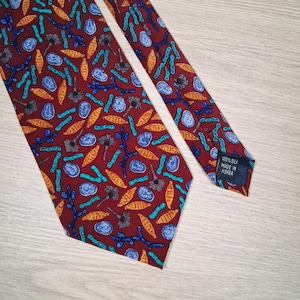 May include: A burgundy silk tie featuring a repeating pattern of teal, orange, and blue abstract shapes. The label reads "100% Silk Made in Korea".