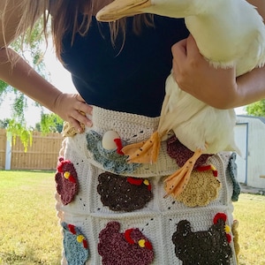 May include: A cream-coloured crochet skirt featuring a grid pattern with various appliqued chickens in shades of brown, red, blue, and yellow. The skirt has a scalloped hem and is worn with a black top, while a white duck is held in front of the skirt.
