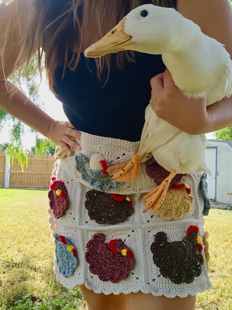 May include: A cream-coloured crochet skirt featuring a grid pattern with various appliqued chickens in shades of brown, red, blue, and yellow. The skirt has a scalloped hem and is worn with a black top, while a white duck is held in front of the skirt.