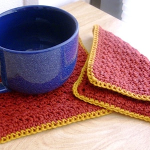 May include: Two crocheted coasters in red and gold. The coasters are square and have a crocheted border. A blue ceramic mug is sitting on one of the coasters.