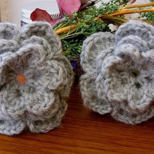 Puede incluir: Dos flores de ganchillo grises con un aspecto arrugado. Las flores están hechas con un hilo suave y peludo y tienen un aspecto delicado y femenino.