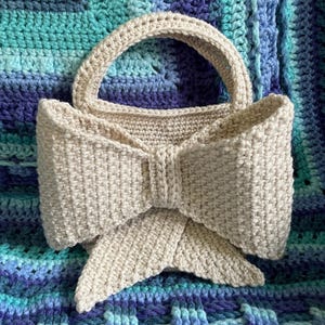 Pu&ograve; includere: Una borsa a mano beige all'uncinetto con un design a fiocco. La borsa ha un manico arrotondato e un grande dettaglio a fiocco sul davanti. La borsa &egrave; appoggiata su una coperta all'uncinetto blu e turchese.