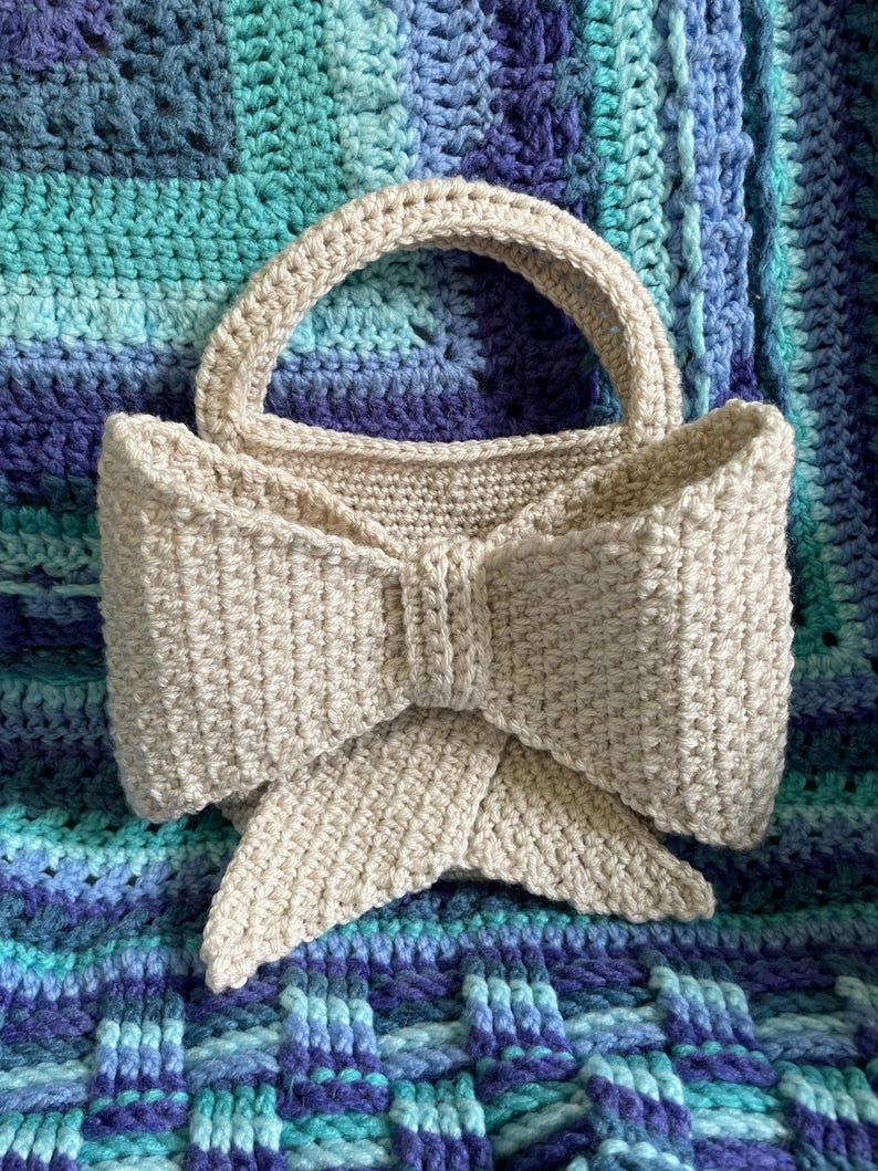 Pu&ograve; includere: Una borsa a mano beige all'uncinetto con un design a fiocco. La borsa ha un manico arrotondato e un grande dettaglio a fiocco sul davanti. La borsa &egrave; appoggiata su una coperta all'uncinetto blu e turchese.