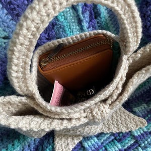 Pu&ograve; includere: Una borsa a mano all'uncinetto beige con manico arrotondato, contenente un portafoglio in pelle marrone, una penna verde e un tubetto rosa. La borsa &egrave; appoggiata su una superficie all'uncinetto blu e viola.