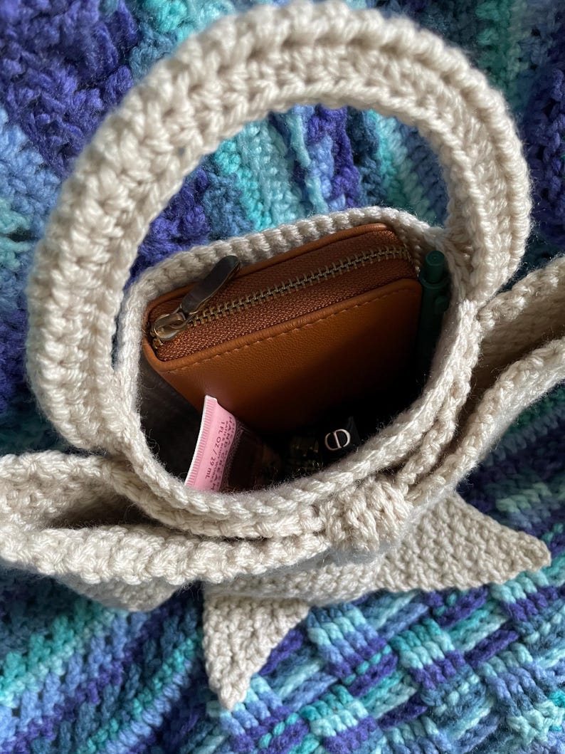 Pu&ograve; includere: Una borsa a mano all'uncinetto beige con manico arrotondato, contenente un portafoglio in pelle marrone, una penna verde e un tubetto rosa. La borsa &egrave; appoggiata su una superficie all'uncinetto blu e viola.