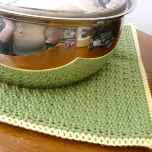 May include: A green crocheted pot holder with a yellow border. The pot holder is on a wooden surface and a silver pot is sitting on top of it.