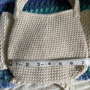 Pu&ograve; includere: Una borsa beige all'uncinetto con manico, di circa 18 cm di larghezza. La borsa &egrave; realizzata con un filato strutturato e poggia su una superficie a fantasia. La forma della borsa &egrave; arrotondata sul fondo.