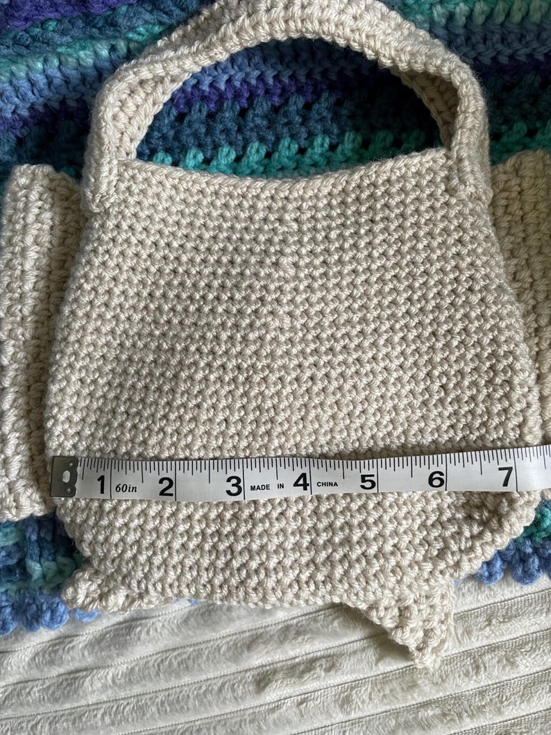 Pu&ograve; includere: Una borsa beige all'uncinetto con manico, di circa 18 cm di larghezza. La borsa &egrave; realizzata con un filato strutturato e poggia su una superficie a fantasia. La forma della borsa &egrave; arrotondata sul fondo.