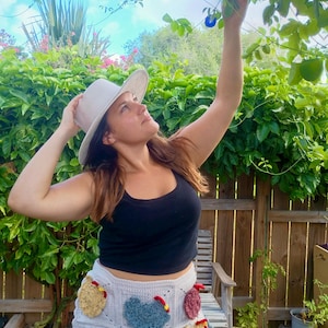 May include: A light grey crochet skirt featuring multiple colourful chicken-shaped appliques in shades of yellow, blue, brown, and red. The skirt is paired with a solid black tank top and a light grey wide-brimmed hat, creating a textured ensemble.