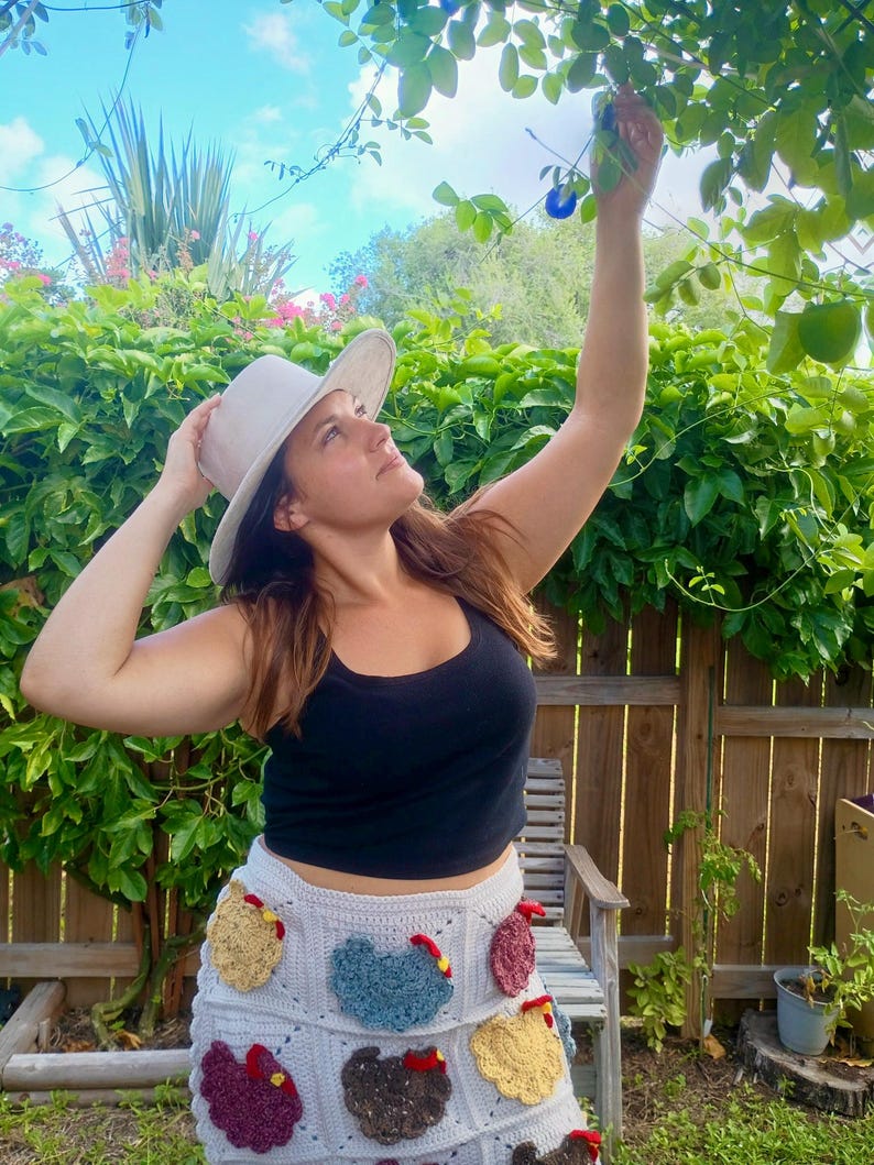 May include: A light grey crochet skirt featuring multiple colourful chicken-shaped appliques in shades of yellow, blue, brown, and red. The skirt is paired with a solid black tank top and a light grey wide-brimmed hat, creating a textured ensemble.