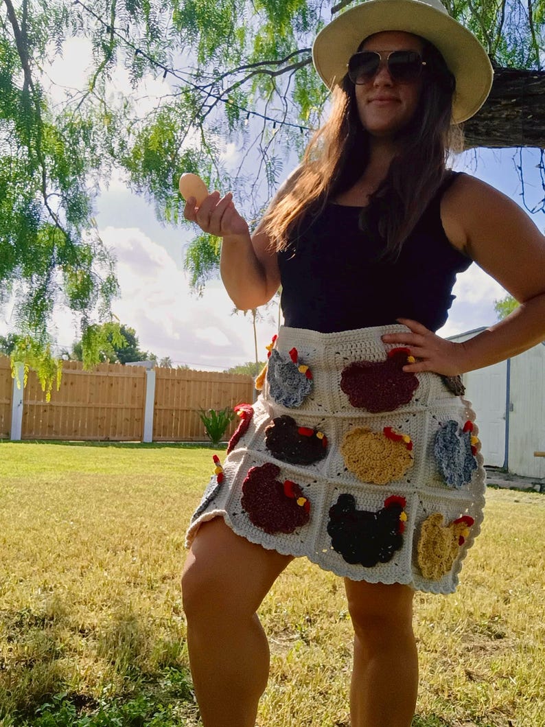 May include: A cream-coloured crochet skirt featuring a grid pattern of individual appliqu&eacute;s shaped like chickens in various colours including black, brown, tan, and blue, each with a small red comb detail, worn with a plain black tank top.