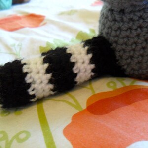 May include: A crocheted raccoon toy with a black and white striped tail. The toy is lying on a white and orange patterned fabric.