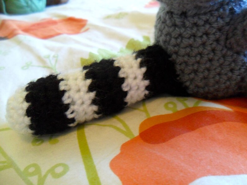 May include: A crocheted raccoon toy with a black and white striped tail. The toy is lying on a white and orange patterned fabric.
