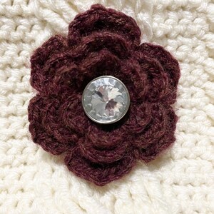 May include: A maroon crocheted flower with a clear rhinestone button in the center, on a white crocheted background.