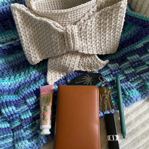 Pu&ograve; includere: Una borsa a mano all'uncinetto beige con un dettaglio a fiocco, appoggiata su una coperta all'uncinetto blu e turchese. Un portafoglio marrone, chiavi, rossetto, penna e crema per le mani sono accanto alla borsa.