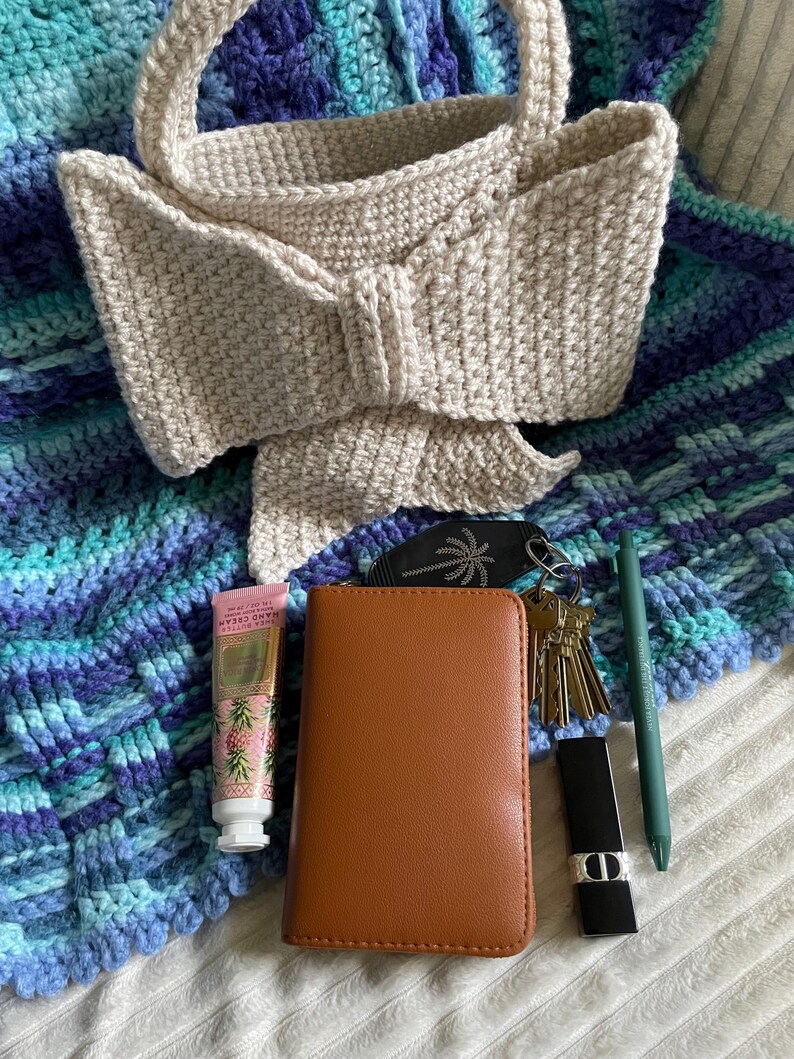 Pu&ograve; includere: Una borsa a mano all'uncinetto beige con un dettaglio a fiocco, appoggiata su una coperta all'uncinetto blu e turchese. Un portafoglio marrone, chiavi, rossetto, penna e crema per le mani sono accanto alla borsa.
