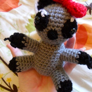 May include: A gray crocheted stuffed animal with black accents and a red bow. The animal has large black eyes and a small black nose.