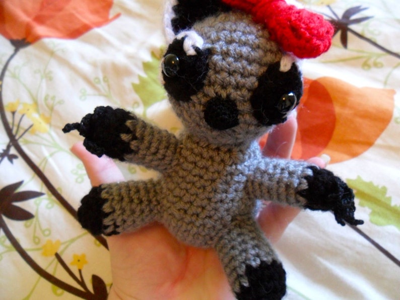 May include: A gray crocheted stuffed animal with black accents and a red bow. The animal has large black eyes and a small black nose.