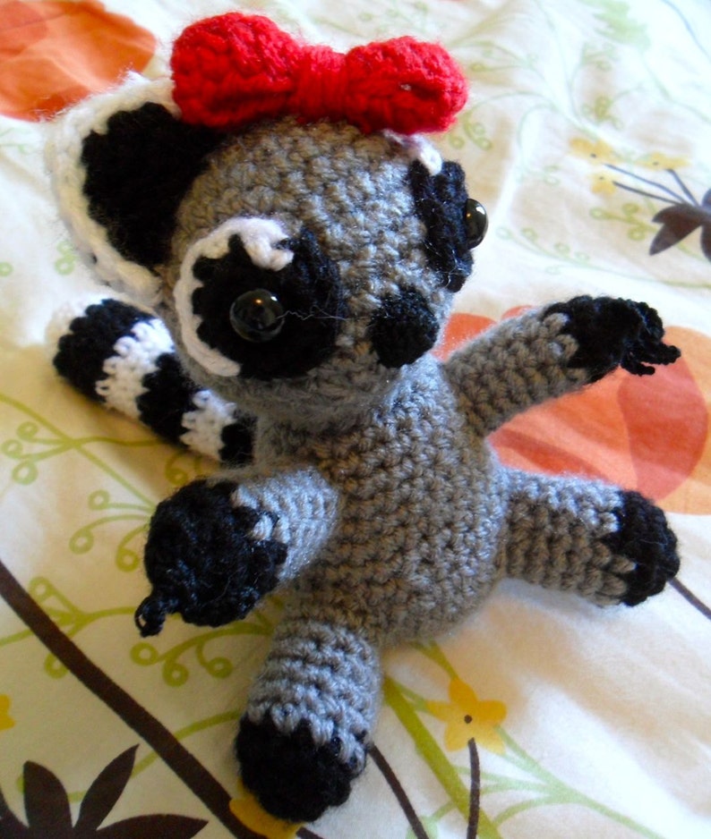 May include: A gray crocheted stuffed animal resembling a lemur with black eyes, a black nose, and a red bow. The animal has white stripes on its arms and legs.