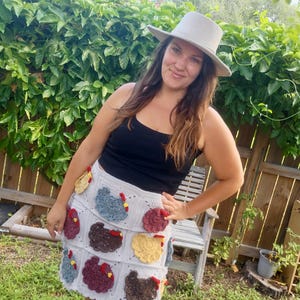 May include: A light grey crochet skirt featuring a grid pattern with various textured chicken appliques in shades of blue, brown, yellow, and maroon. The skirt has a scalloped hem and is paired with a simple black tank top and a light grey fedora hat.