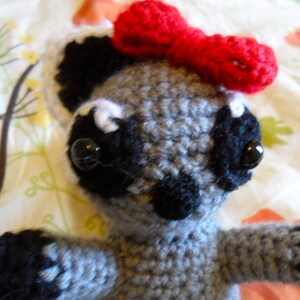 May include: A gray crocheted raccoon with black eyes and a red bow. The raccoon is sitting on a white and green floral fabric.