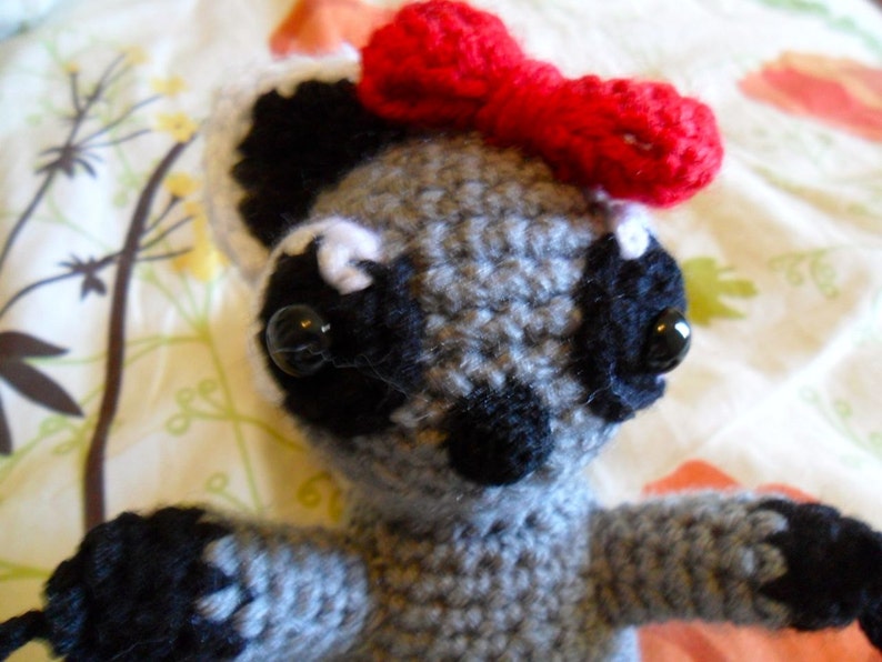 May include: A gray crocheted raccoon with black eyes and a red bow. The raccoon is sitting on a white and green floral fabric.
