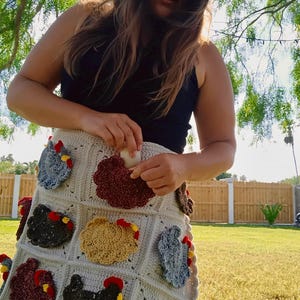 May include: A cream-coloured crochet apron featuring multiple pockets, each adorned with a textured, multi-coloured chicken motif. The chickens are crafted in shades of brown, grey, and gold, with red combs and yellow beaks, integrated into a grid pattern.