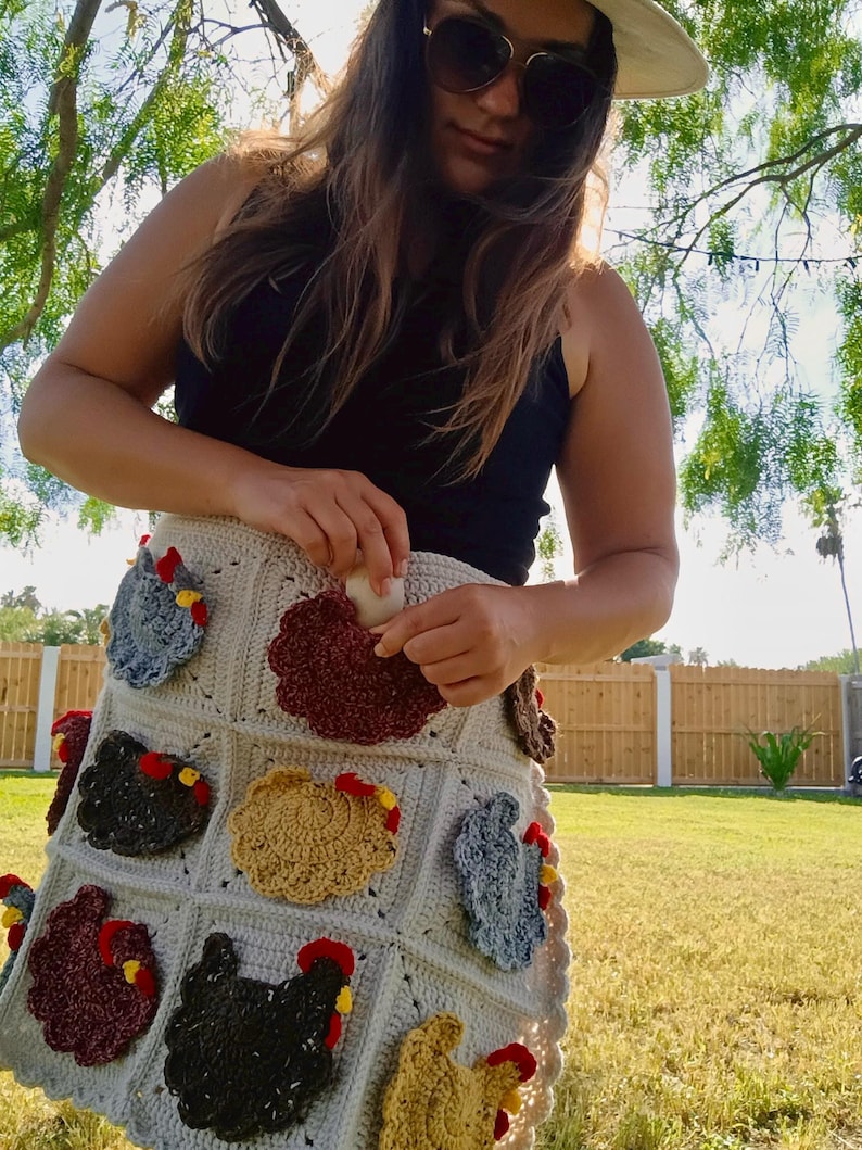 May include: A cream-coloured crochet apron featuring multiple pockets, each adorned with a textured, multi-coloured chicken motif. The chickens are crafted in shades of brown, grey, and gold, with red combs and yellow beaks, integrated into a grid pattern.