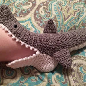 May include: Grey and white crocheted shark slipper with a white crocheted trim around the mouth. The slipper is on a patterned fabric.