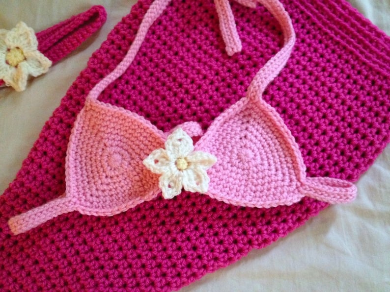 May include: A pink crocheted mermaid tail with a matching pink and white crocheted top and a white flower accent.