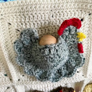 May include: A cream-coloured crocheted square featuring a three-dimensional grey chicken appliqu&eacute; with a red comb, a yellow beak, and a red wattle. A smooth, round, light-coloured wooden bead is nestled within the centre of the grey crocheted chicken.