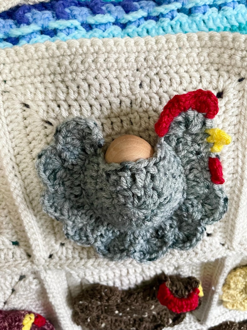 May include: A cream-coloured crocheted square featuring a three-dimensional grey chicken appliqu&eacute; with a red comb, a yellow beak, and a red wattle. A smooth, round, light-coloured wooden bead is nestled within the centre of the grey crocheted chicken.