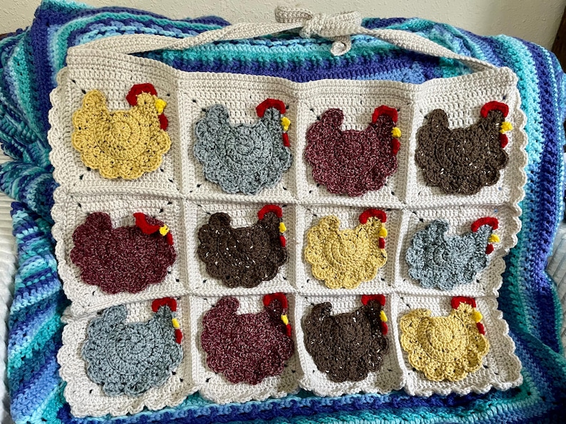 May include: A cream-coloured crocheted wall hanging featuring twelve square panels, each decorated with a textured chicken applique in various colours including yellow, grey, maroon, and brown, each with a small red and yellow comb detail.
