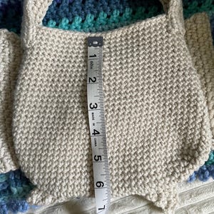 Pu&ograve; includere: Una borsa all'uncinetto fatta a mano color crema con un manico. La borsa &egrave; alta circa 15 cm. La borsa &egrave; appoggiata su una coperta all'uncinetto blu e verde.