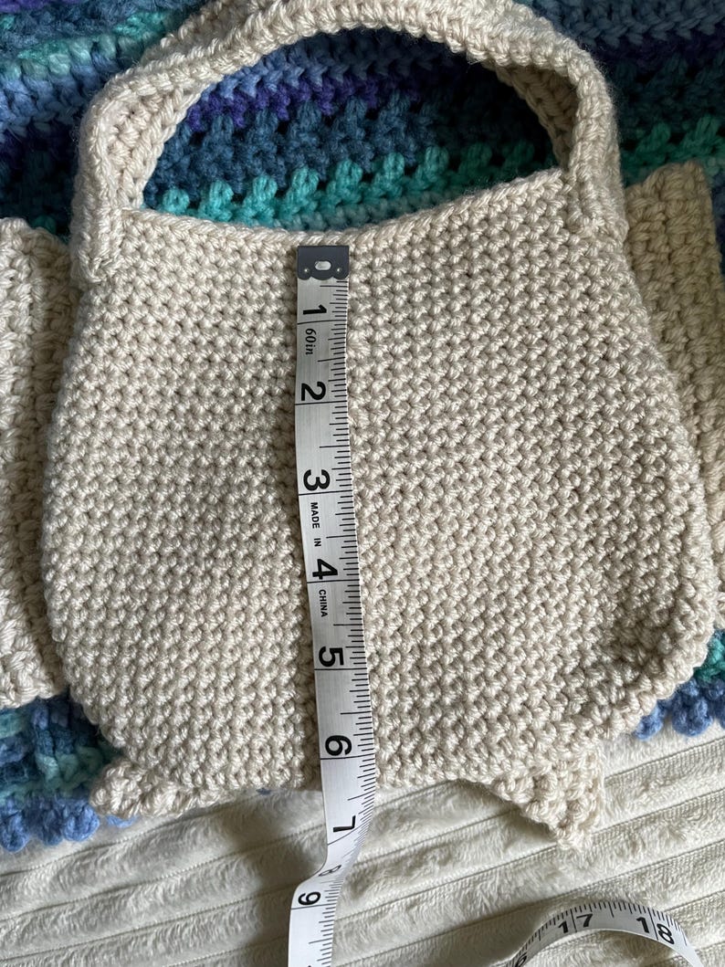 Pu&ograve; includere: Una borsa all'uncinetto fatta a mano color crema con un manico. La borsa &egrave; alta circa 15 cm. La borsa &egrave; appoggiata su una coperta all'uncinetto blu e verde.