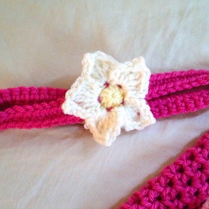 May include: A pink crocheted headband with a white crocheted flower accent. The flower has a yellow center.