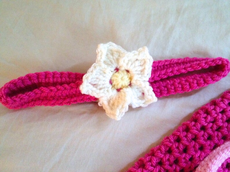 May include: A pink crocheted headband with a white crocheted flower accent. The flower has a yellow center.