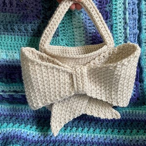 Pu&ograve; includere: Una borsa all'uncinetto beige con un davanti a forma di fiocco. La borsa ha un corpo rettangolare e due manici arrotondati. Lo sfondo &egrave; una coperta all'uncinetto blu e turchese.