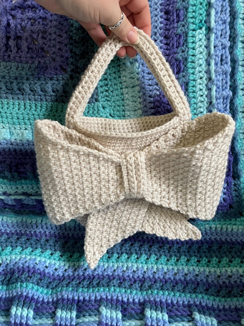Pu&ograve; includere: Una borsa all'uncinetto beige con un davanti a forma di fiocco. La borsa ha un corpo rettangolare e due manici arrotondati. Lo sfondo &egrave; una coperta all'uncinetto blu e turchese.