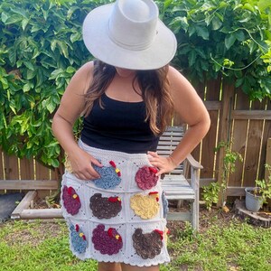 May include: A light grey crochet skirt featuring a grid pattern of individual squares, each decorated with a textured, multi-coloured chicken appliqu&eacute; in shades of blue, yellow, brown, and maroon, complete with small red combs and yellow beaks.