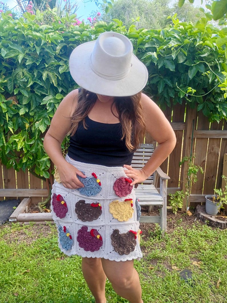 May include: A light grey crochet skirt featuring a grid pattern of individual squares, each decorated with a textured, multi-coloured chicken appliqu&eacute; in shades of blue, yellow, brown, and maroon, complete with small red combs and yellow beaks.